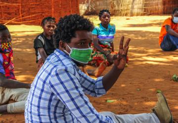 Un hombre en el centro de la imagen con una mascarilla muestra tres dedos de su mano, mientras otras personas socialmente distanciadas se sientan en un semicírculo al fondo.