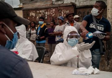 La imagen muestra a personas esperando detrás de una mesa de trabajadores de la salud, quienes usan equipo de protección personal de pies a cabeza. Un hombre parado detrás de los trabajadores de la salud vierte desinfectante en las manos de uno de los trabajadores, mientras que otro hombre (que vemos de espaldas) los observa y espera.