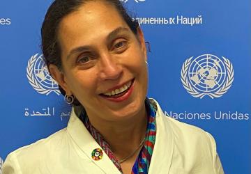 A woman in a yellow suit smiles at the camera in front of a blue and white United Nations sign.