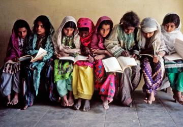 Varios niños y niñas vestidos con colores vivos miran con atención el contenido de los los libros que tiene sobre el regazo.