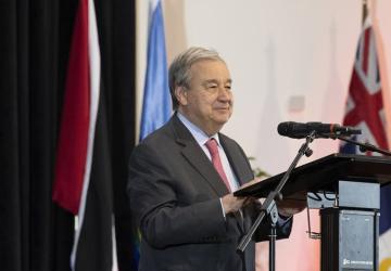 رجل يرتدي بدلة وربطة عنق حمراء، يقف ويتحدث أمام ميكروفون على منصة، وتحيط به مجموعة من الأعلام.