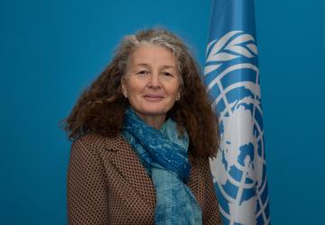 A woman in a brown blazer and blue scarf stands next to a UN flag.