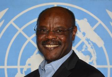 man in dark suit stands in front of UN flag 