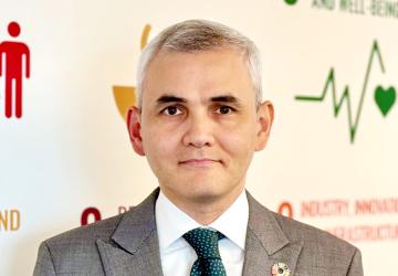 A man in a grey suit and green tie stands in front of a wall painted with the colours of the SDGs