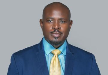 A man in a blue shirt, blue blazer and yellow tie against a grey background.