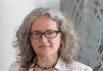 Une femme aux cheveux argentés et aux lunettes foncées se tient devant un mur de verre arborant le logo de l'ONU.