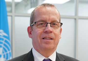 Un homme portant des lunettes et un costume sombre se tient devant un drapeau bleu portant le logo de l'ONU.