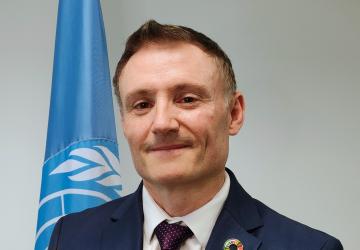 A man in a blue suit stands in front of a blue UN flag