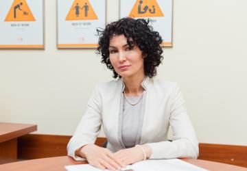 A woman in a white suit and black curly hair sits at a brown wooden desk with her arms crossed