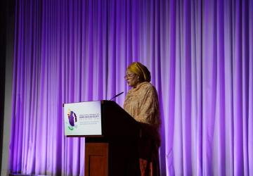 A woman stands in front of a purple background. She is speaking at a lectern and the sign on the lecturn reads Women, Peace and Security conference
