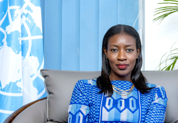 A woman in a blue dress sits on a grey chair against a blue UN flag