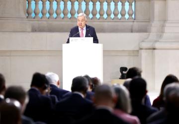 A man in a dark suit speaks into a microphone at a podium while several other people in silhouettes listen on