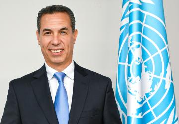 A man in a black suit and blue tie stands and smiles in front of white wall with a blue flag, the UN flag.