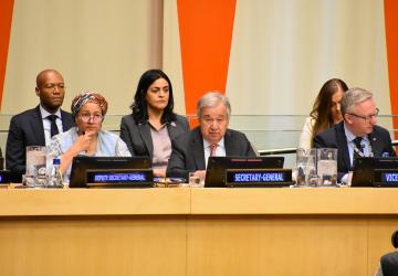 A group of 3 people at a podium in the United Nations. Behind them are three people sitting in chairs.