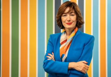 A woman in a blue suit and orange coloured scarf stands with her arms folded against a colourful wall.