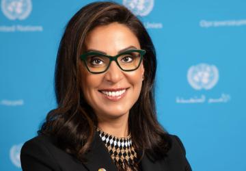 The RC is looking at the camera and smiling. She is wearing a black jacket and glasses and standing in front of a UN blue background with the UN logo imprinted