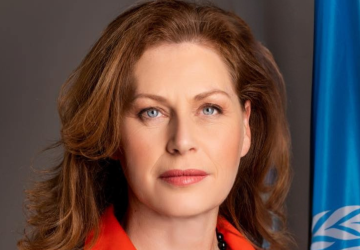 A woman with brown hair and a red shirt stands next to a blue UN flag