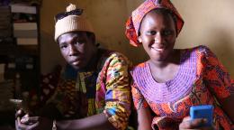 Une femme et un homme sénégalais vêtus d'habits traditionnels sont assis côte à côte et regardent l'objectif de la caméra en souriant et en tenant leur téléphone portable dans les mains.