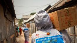 Une personne, dos à la caméra, marche le long d’un sentier séparant deux rangées de cabanes. Elle porte un carton sur l’épaule droite et est vêtue d’un T-shirt sur lequel figurent des inscriptions en bengali et le logo d’UK Aid.