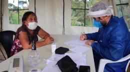 Un homme et une femme sont assis autour d’une table dans ce qu’on devine être une très grande tente. L’homme, à droite de l’image, est un membre du personnel de l'ONU. Il porte un masque, une visière et une tenue de protection bleue et il remplit un document administratif. La femme assise devant lui, à gauche de l’image, patiente en regardant la caméra.