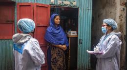 Tres mujeres se paran frente a la fachada de una tienda a pie de calle. Una de las personas es una residente local. Lleva un vestido y una gran estola azul, alrededor de cabeza, hombros y torso. Las otras dos personas usan mascarilla y equipo de protección personal. Una de las personas con equipo de protección está tomando notas sobre una tablilla con sujetapapeles.
