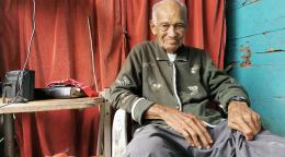 Candelario, un homme âgé, est assis près d'une table sur laquelle se trouve du matériel électronique. Il regarde la caméra en souriant.