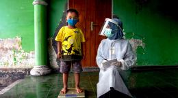 طفل صغير يقف على الميزان وهو ينظر إلى الكاميرا مرتديًا كمامة، ويركع عامل رعاية صحية بجانبه.