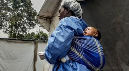 Una mujer vestida con equipo de protección personal carga a un bebé en la espalda.