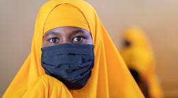 A young adolescent girl wearing a yellow headdress and black mask looks strikingly at the camera.