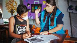 A teacher in a blue vest and face mask talks to a boy in a black tank top and white face mask. 
