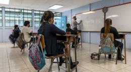 Children seated at dDes écoliers et écolières portant un masque de protection respiratoire sont assis dans une salle de classe et suivent le cours d’un enseignant que l’on voit écrire sur un tableau blanc.esks attending in-person school at a social distance wearing masks. 