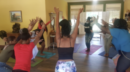 Several women, in a yellow room, have their hands while practicing yoga in a group.