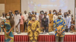 Un gran grupo de personas con máscaras faciales se sitúan frente a la cámara, delante de un proyector que muestra las palabras "Hack COVID-19".