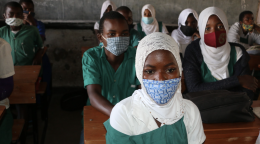 A group of children in school uniforms and masks sit in a classroom facing forward.