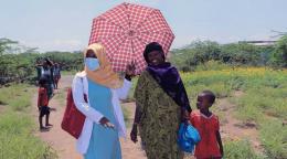 One woman wearing a face masks holds an umbrella as she walks side-by-side with a woman and her child.