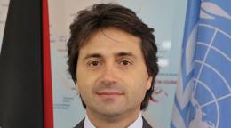 A man in a brown tie and jacket smiles straight at the camera in front of two flags.