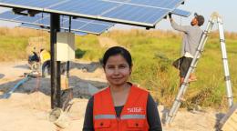 A woman in an orange vest smiles in front of large solar panels. 