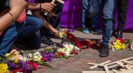 Image shows the people's arms holding crosses kneeled by flowers and crosses piled in front of them. 