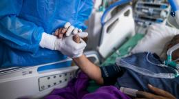 A patient and a medical professional hold hands as the person lies in a hospital bed. 
