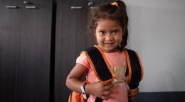 Una niña con una pequeña sonrisa y que mira estoicamente hacia la cámara mientras se agarra a las correas de su mochila.
