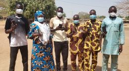 Un groupe de personnes portant un masque de protection respiratoire se tiennent debout, en extérieur, et montrent leur téléphone portable à la caméra.