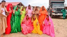 Un grupo de personas vestidos de colores se sitúan en un semicírculo sobre un fondo arenoso de color tostado.