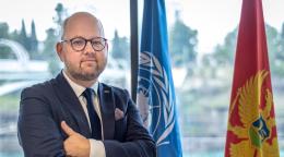 Un hombre con traje junto a las banderas de Naciones Unidas y Montenegro.