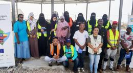 Un groupe d’hommes et de femmes, dont certaines portent un voile intégral noir, se tiennent côte à côte, en extérieur, et regardent en direction de la caméra en souriant.