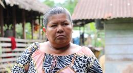 Plano detalle de una mujer indígena que mira con rostro serio y determinación a la cámara. Se encuentra en un espacio abierto, con vegetación tropical y dos estructuras rústicas, tradicionales de la zona y semi techadas a manera de fondo.
