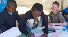En Madagascar, dos niños estudian en una mesa bajo una gran tienda de campaña instalada por UNICEF, mientras un tercer niño, albino, les observa con una gran sonrisa.