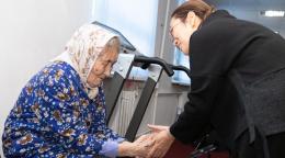 Older woman on the left in blue dress and headscarf holding hands with woman on the right in a black dress