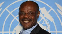 man in dark suit stands in front of UN flag 