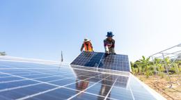 Two people in hats and colourful clothes stand on one side of a silver coloured solar panel in Cambodia