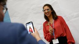 A woman in a brown shirt with glasses shaped like the UN SDGs smiles as someone else takes a photo of her on their mobile phone